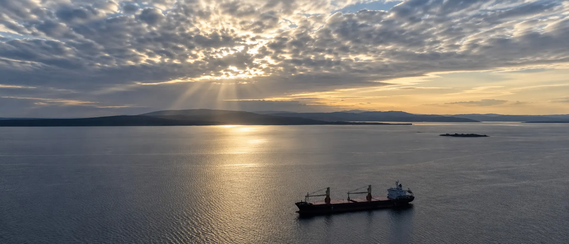 A geared bulk carrier lying at anchor at sunset, low hills across the water behind her.