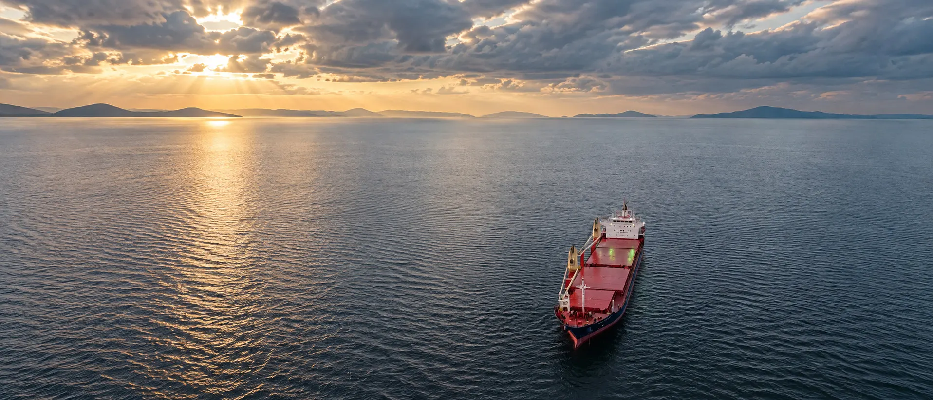 A bulk carrier under way at dusk on open water, sunbeams breaking through cloud over hills on the horizon.