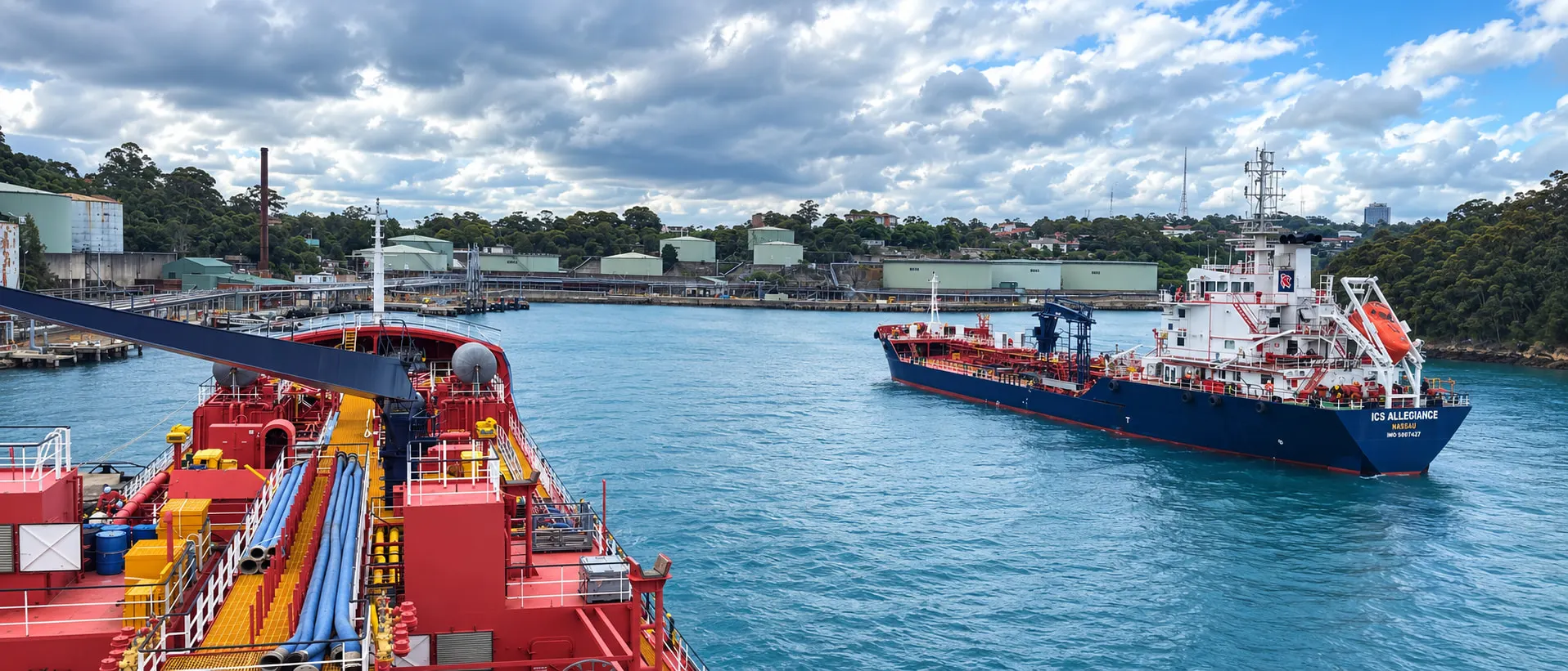 Looking across a harbour from a tanker’s deck to a bunker vessel under way, with a fuel terminal and wooded shore behind her.