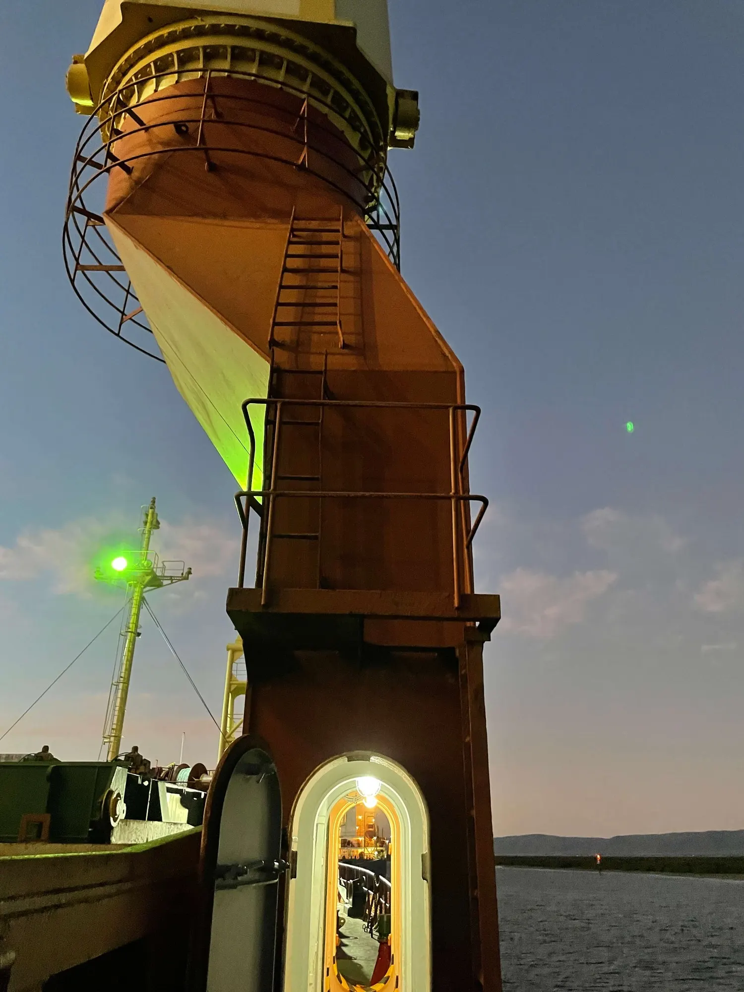 A deck crane and its lit access door at dusk, the green starboard navigation light burning on the mast beyond.