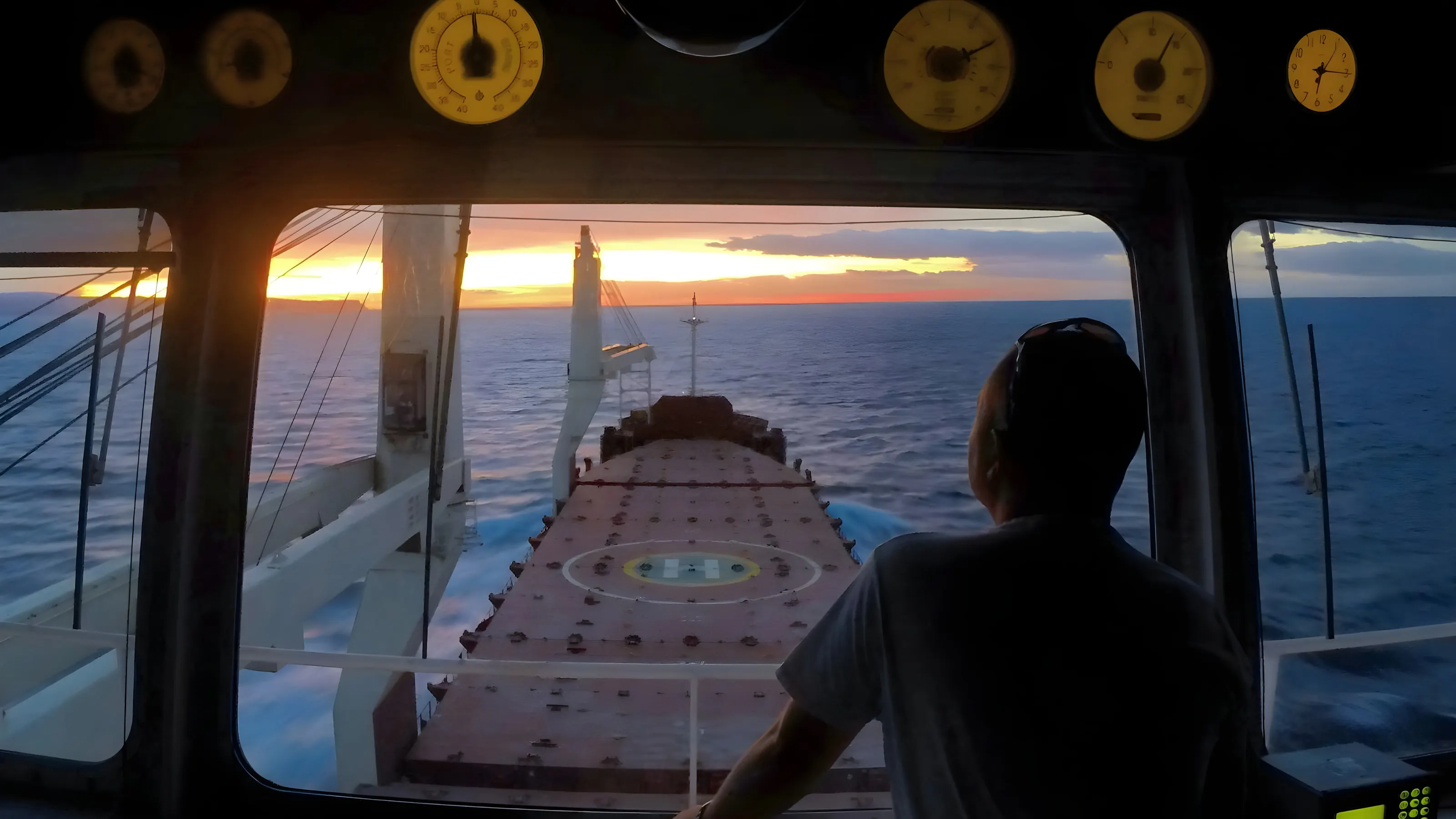 The view forward from a ship’s bridge at sunset, an officer at the console watching the foredeck.