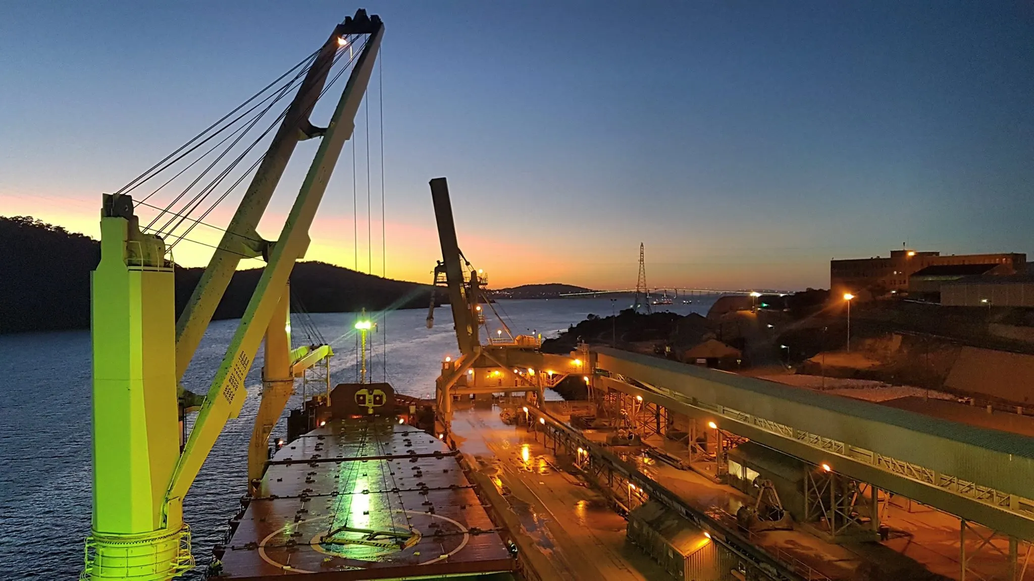 A geared bulk carrier alongside at dusk, her cranes raised over the hatches and the shore loader working beside her.