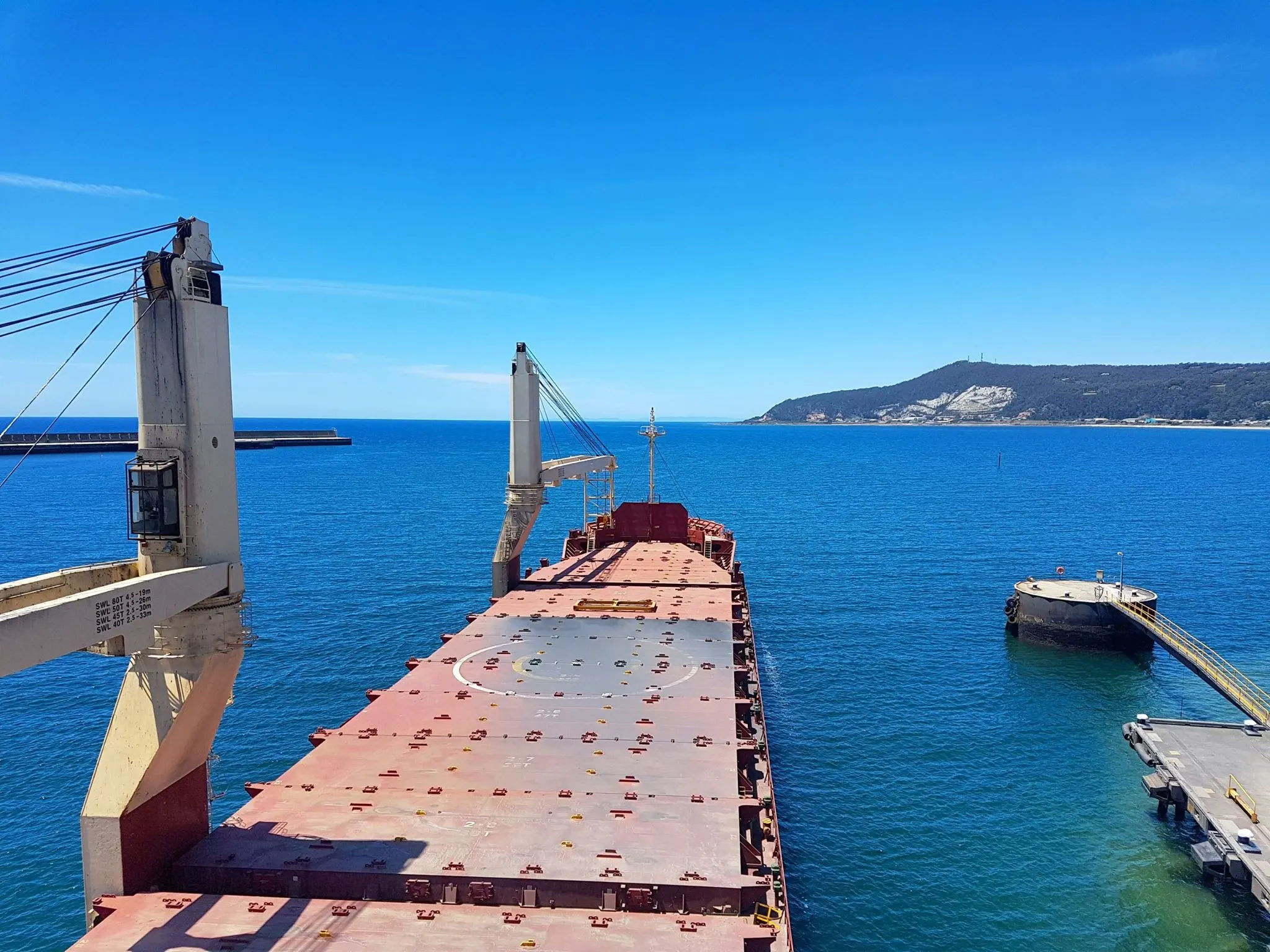Looking forward along a loaded cargo deck to a headland, a deck crane standing over the hatch covers.