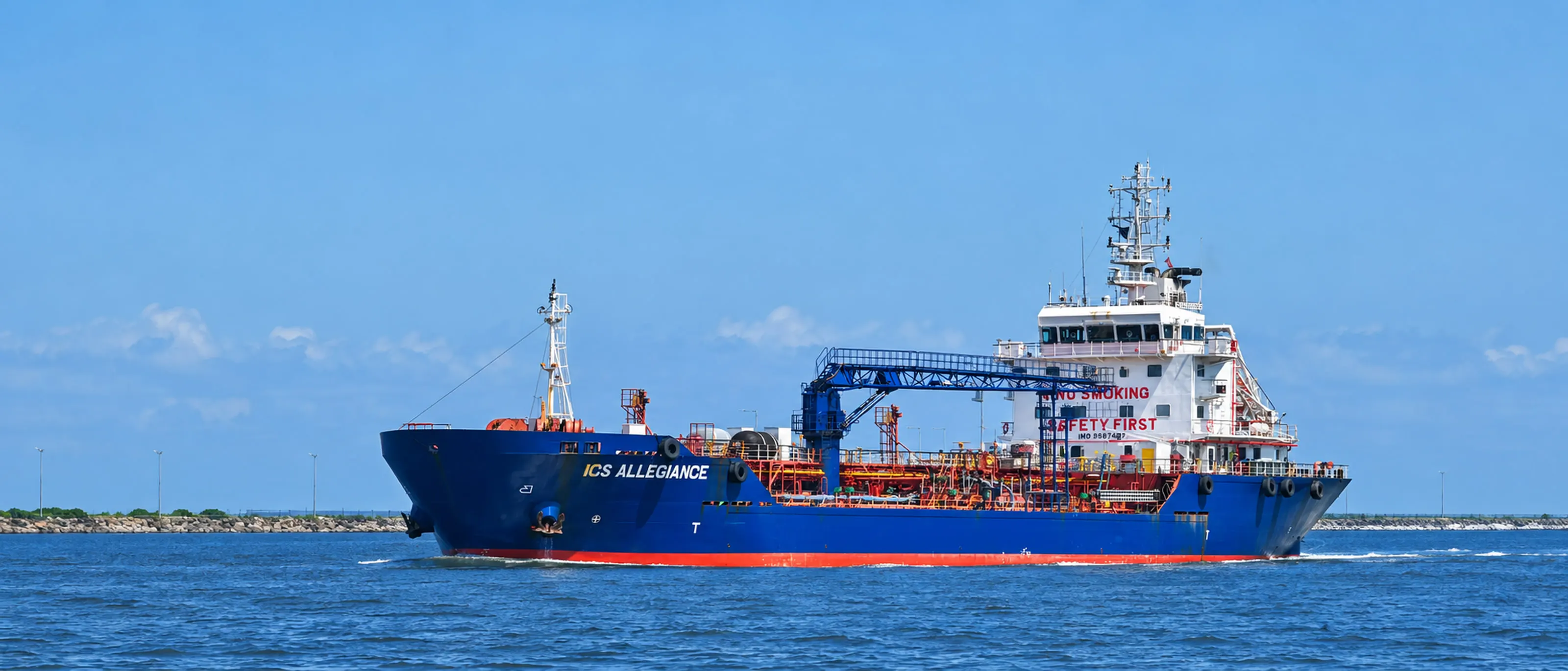 ICS Allegiance under way in open water, port side on, with a long breakwater and a clear sky behind her.