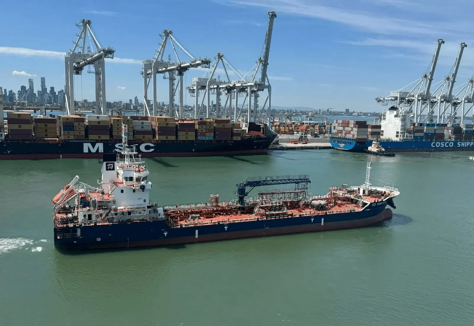 ICS Tenacious alongside at a container terminal, shore gantries and a city skyline behind her.