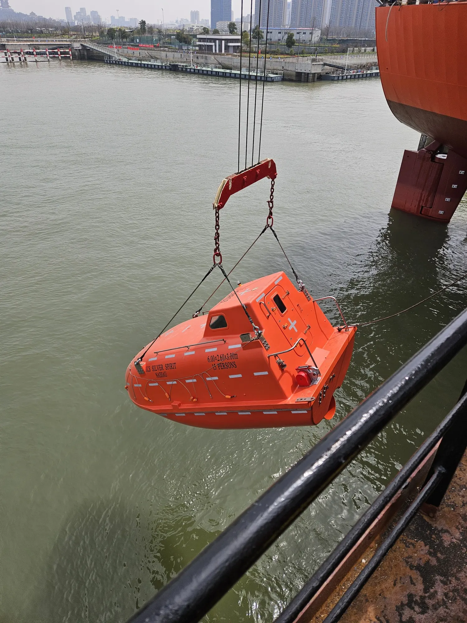 The enclosed lifeboat hanging in its falls above the water alongside, a city waterfront beyond.