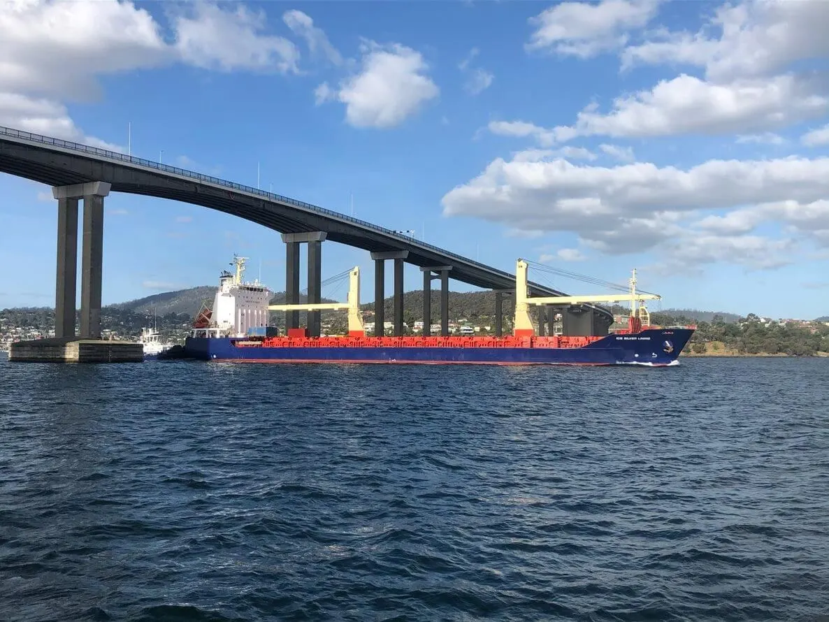 ICS Silver Lining in ballast passing beneath a long road bridge, hills behind her.