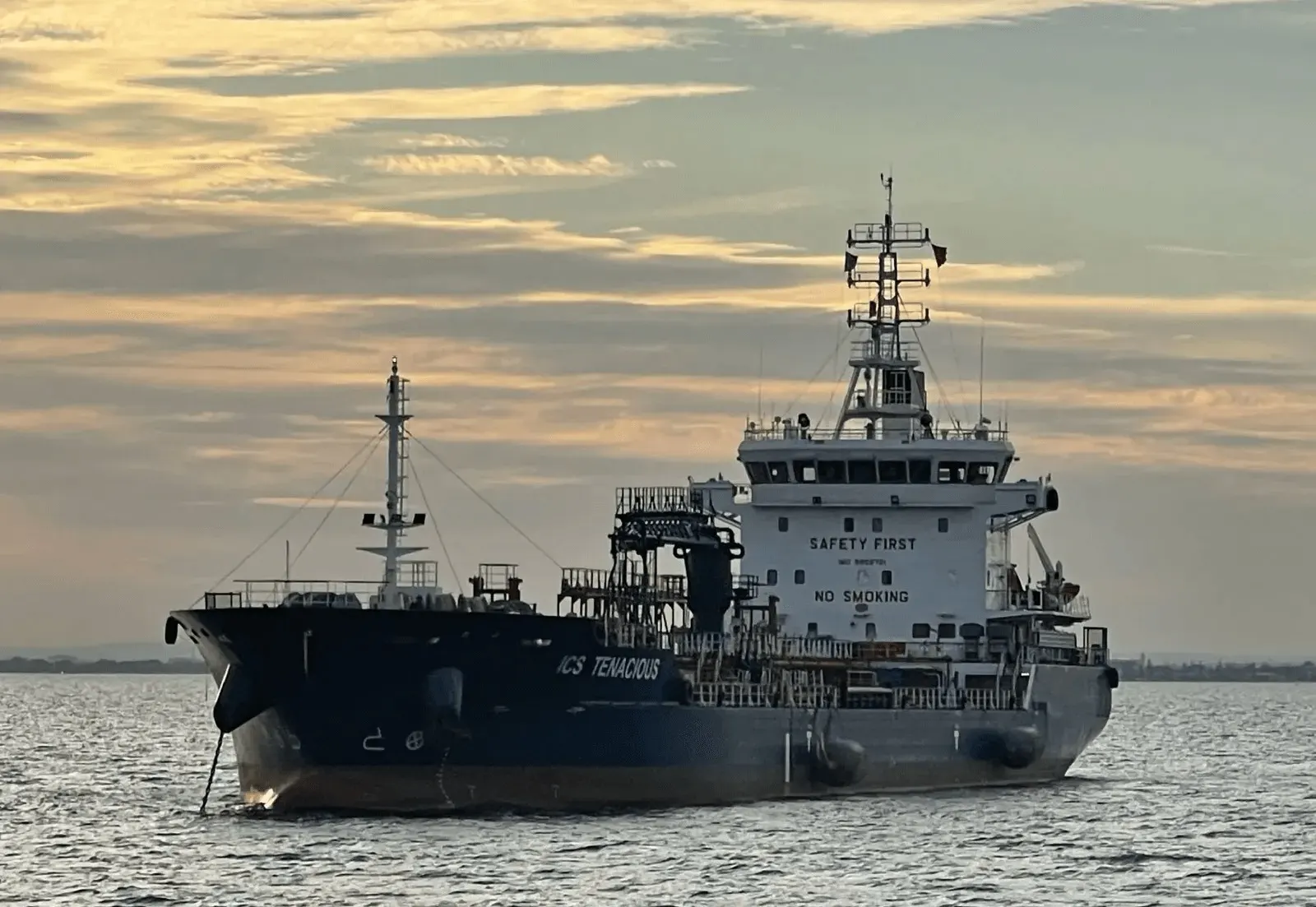 ICS Tenacious lying still in flat water at first light, her hull dark against a pale sky.