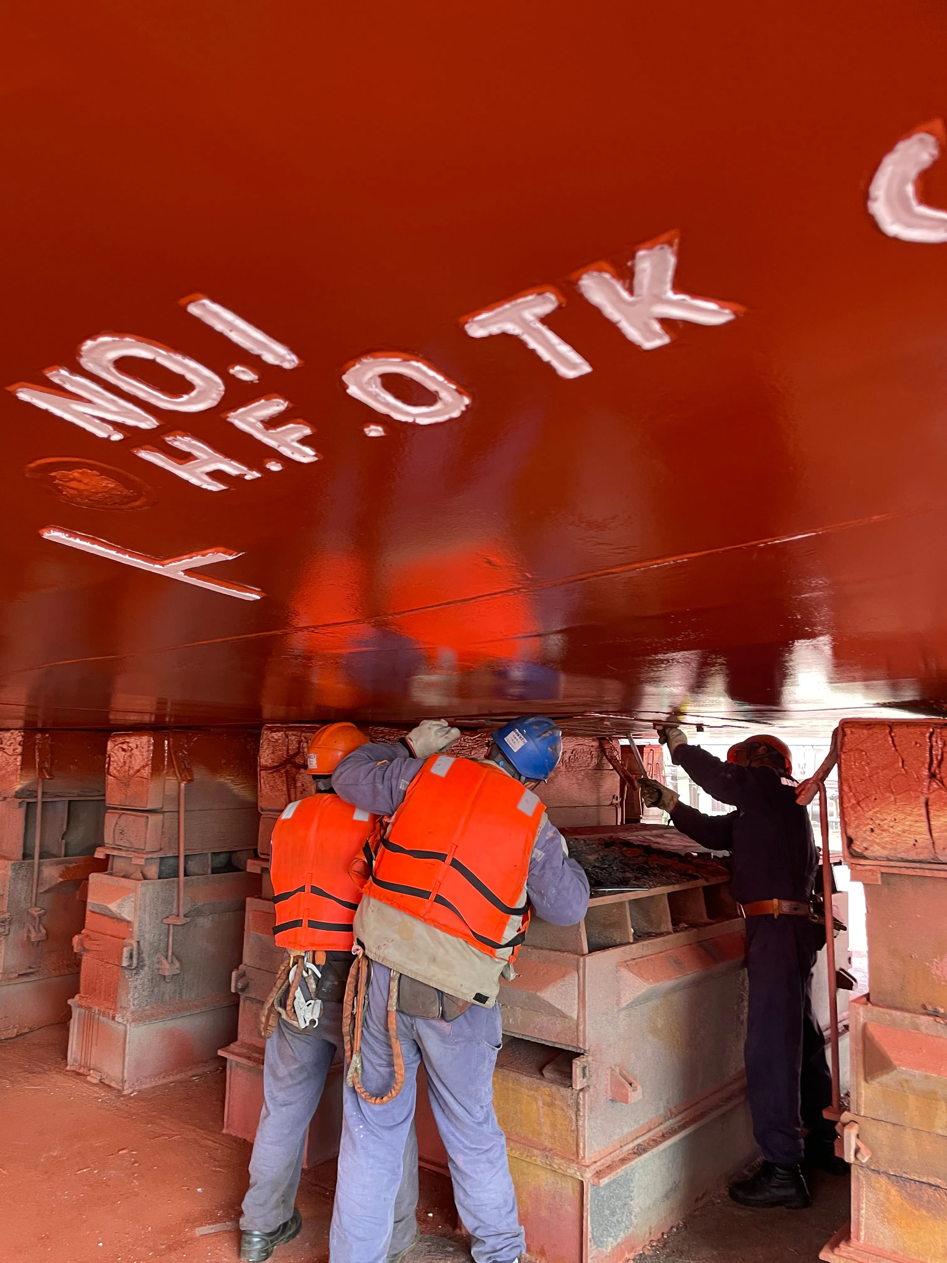 Workers under a freshly painted hull in dry dock, beside the keel blocks.