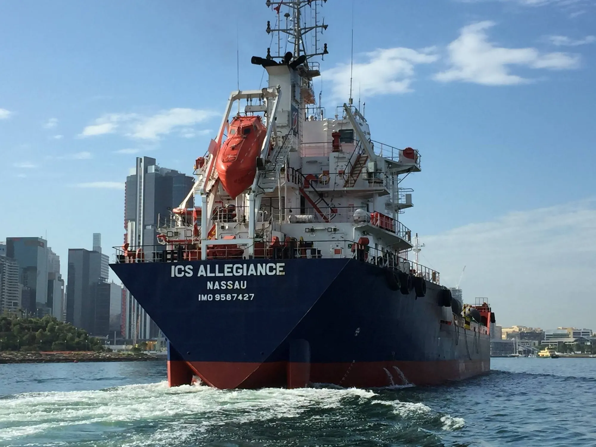 ICS Allegiance under way bow-on with a city skyline behind her and her bow wave turning over.